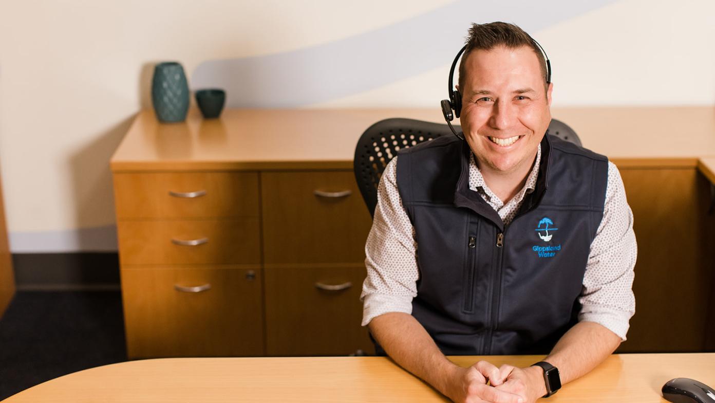A male Gippsland Water team member is sitting at a desk wearing a headset. His hands are held together and he's smiling at the camera.