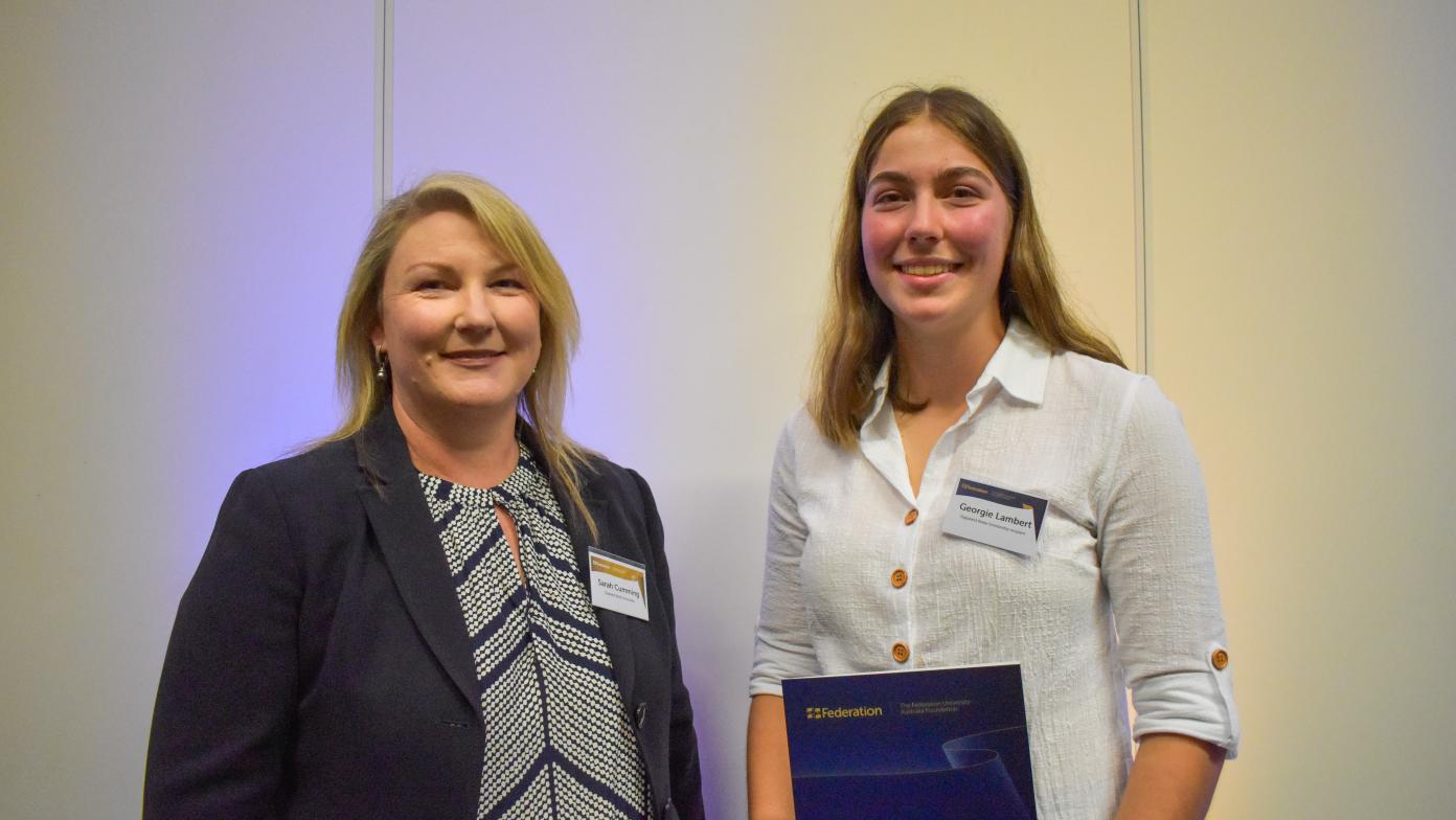 Managing Director Sarah cumming and 2022 scholarship recipient Georgie Lambert are standing next to each other. Sarah is on the left, and Georgie is on the right. They are both looking at the camera smiling, and Georgie is holding her scholarship. 