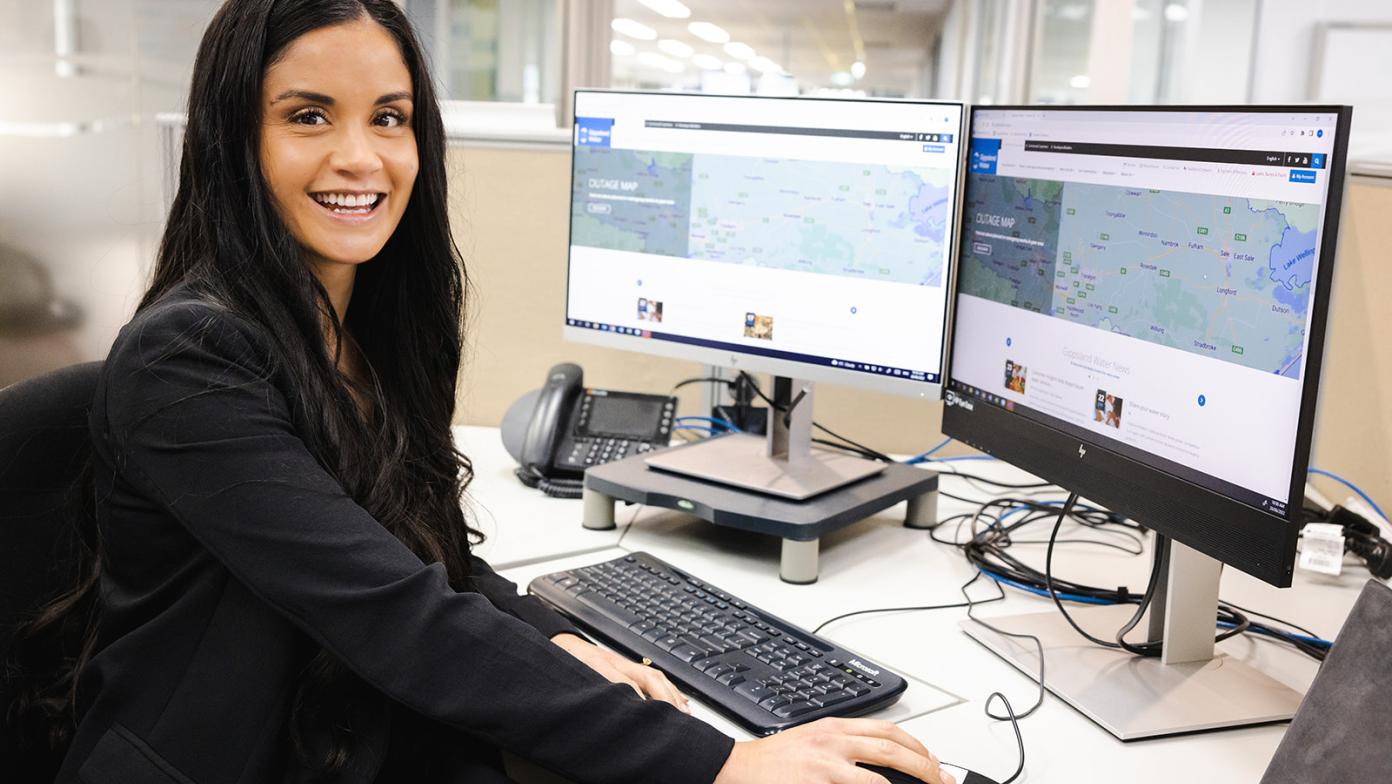 Gippsland Water employee sits at desk with two computer screens displaying website. Employee is looking at the camera and smiling.