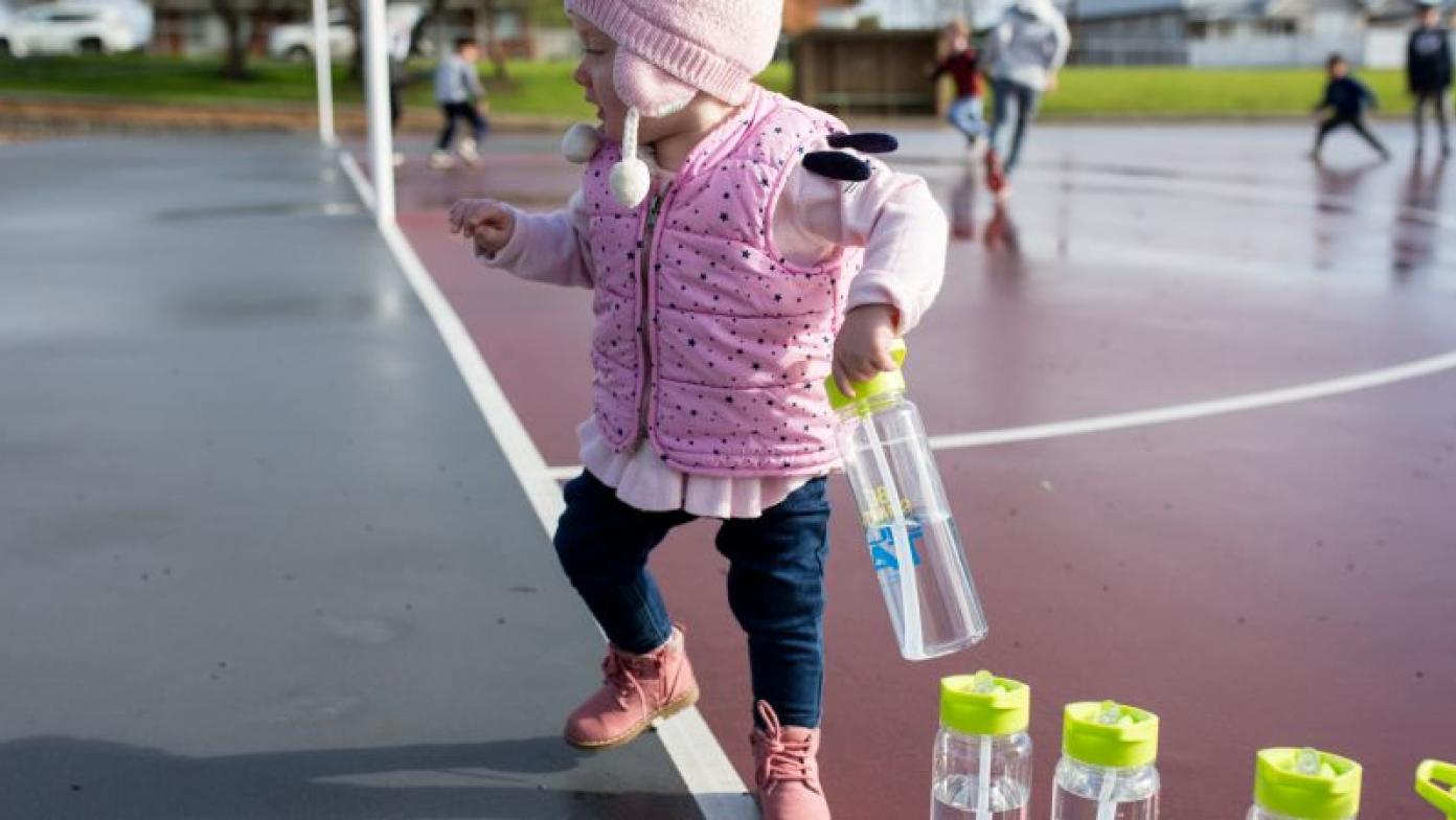Toddler runs around netball court with clear drink bottle. 