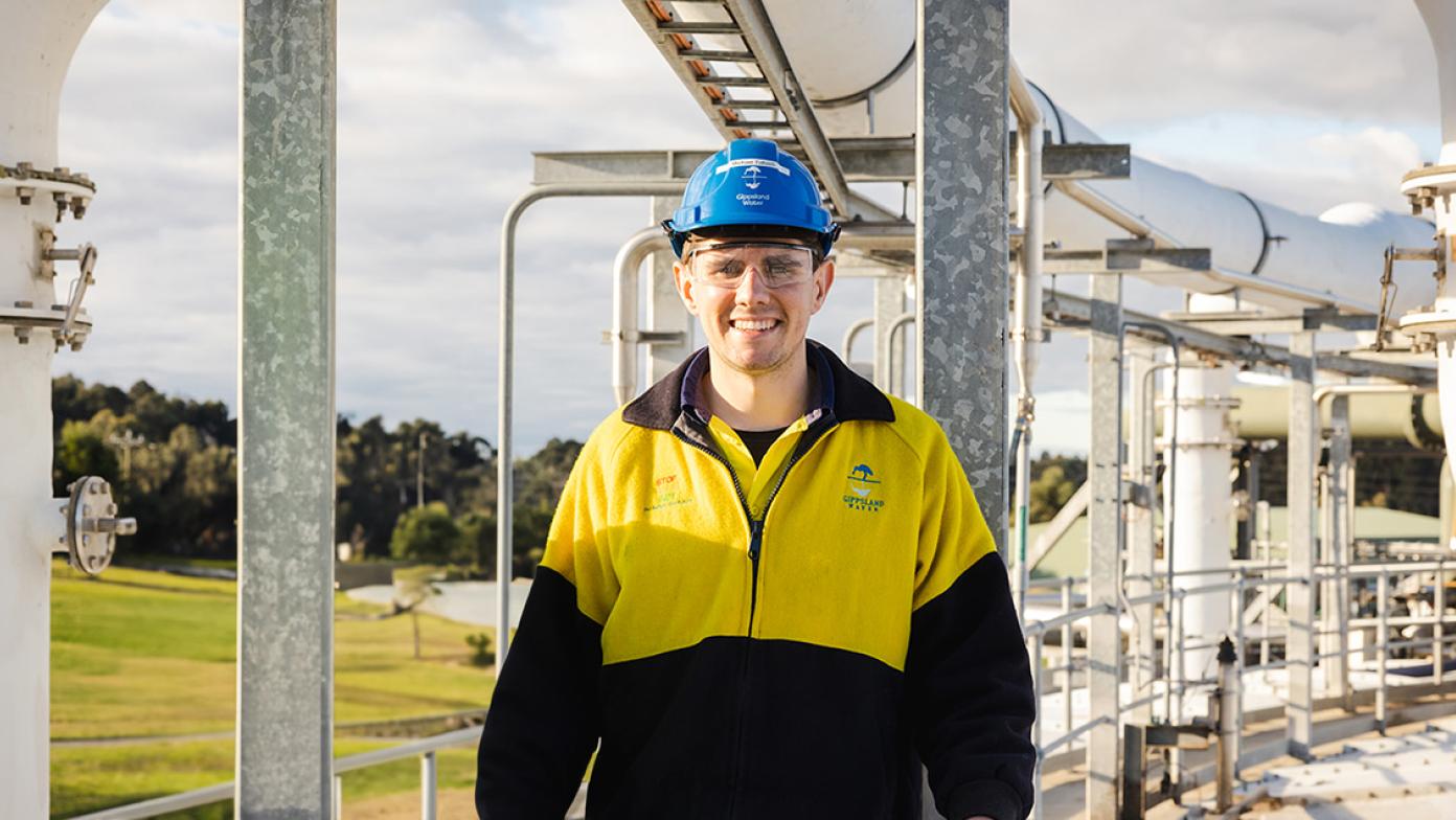 Michael is at the water factory wearing hi-vis and a hard hat. He's smiling and looking at the camera.