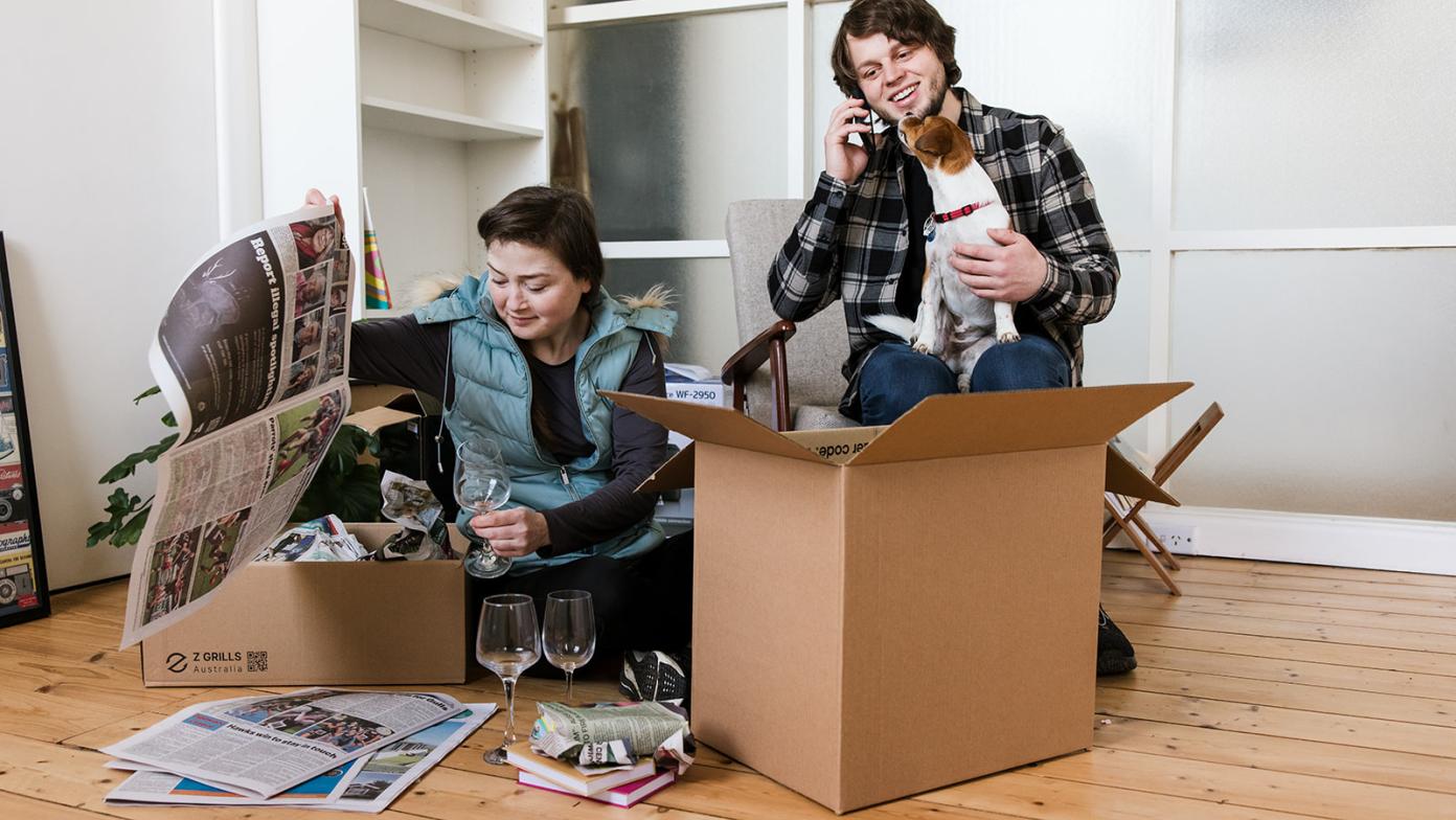 A couple moving house. A woman is packing a box on the floor, a man is on the phone.