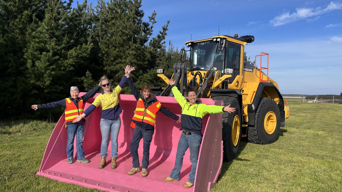 Three people stand in a pink bucket with their arms in the air. 