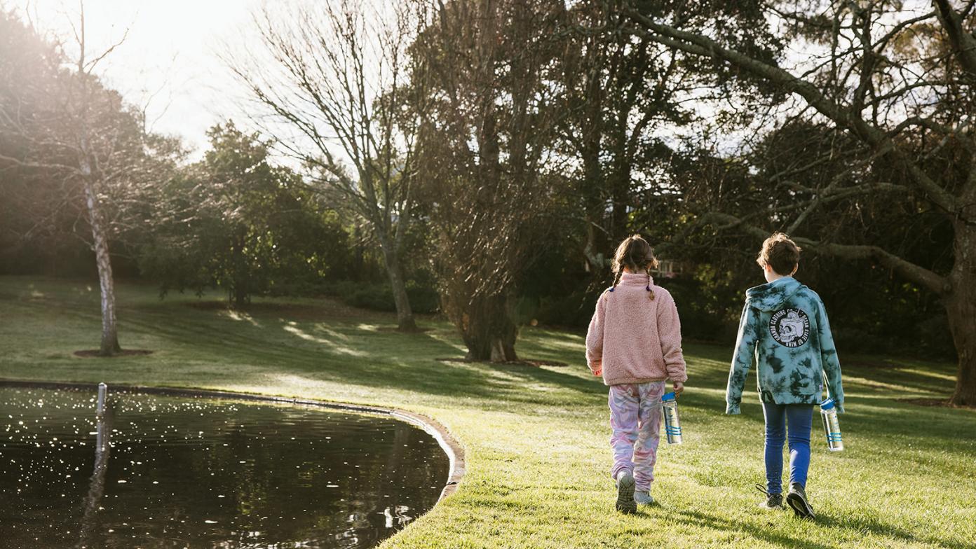 Two children walking on a grass field next to a lake.