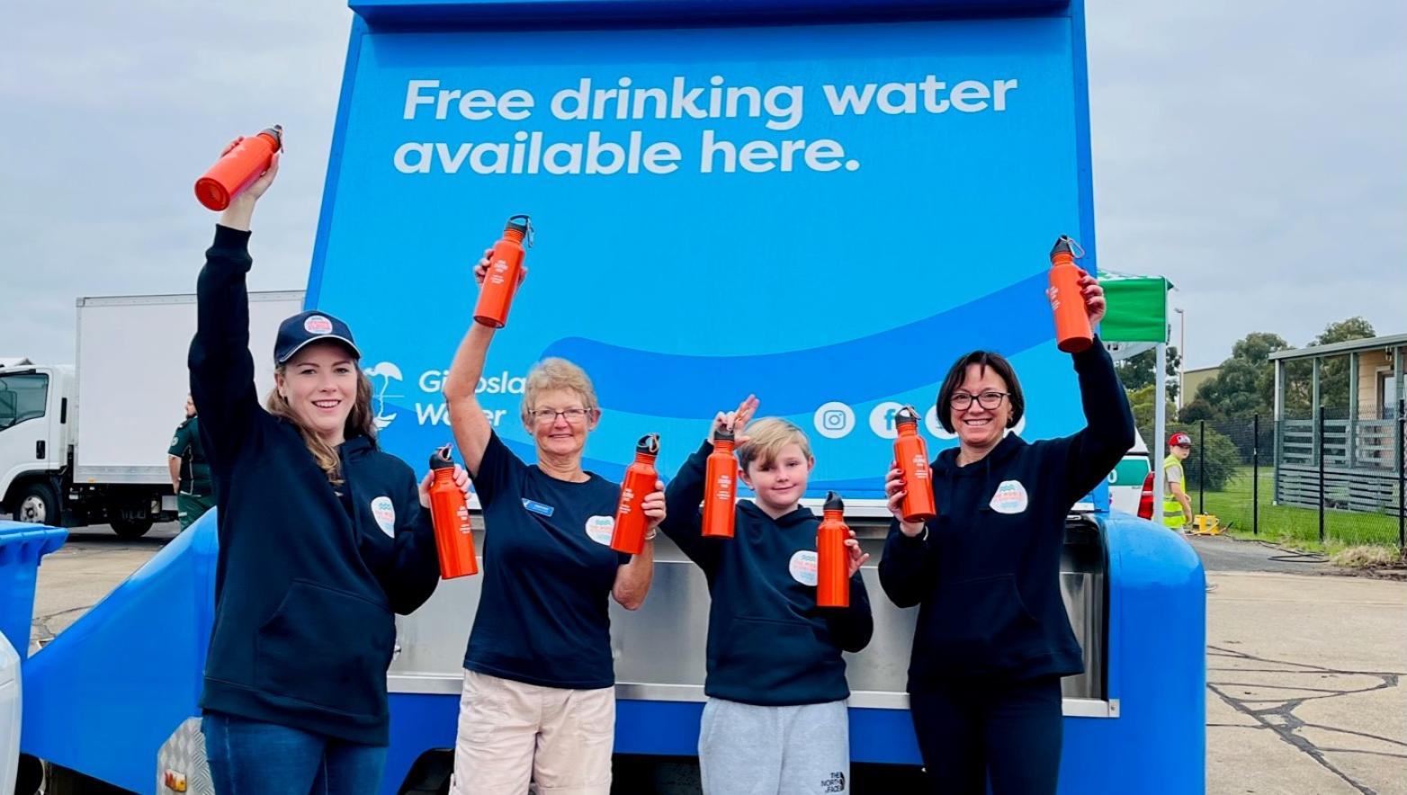 Four customers are standing in front of a large blue water trailer. The customers are wearing navy uniform and holding red drink bottles above their heads. The water trailer is taller than the customers and has a sign saying ‘Free drinking water available here’. 