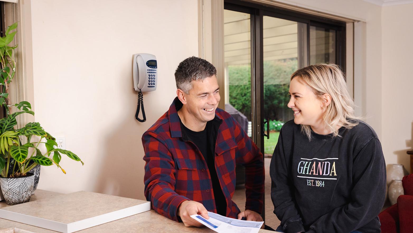 Man and woman sit at kitchen bench smiling. Man holds water bill. 