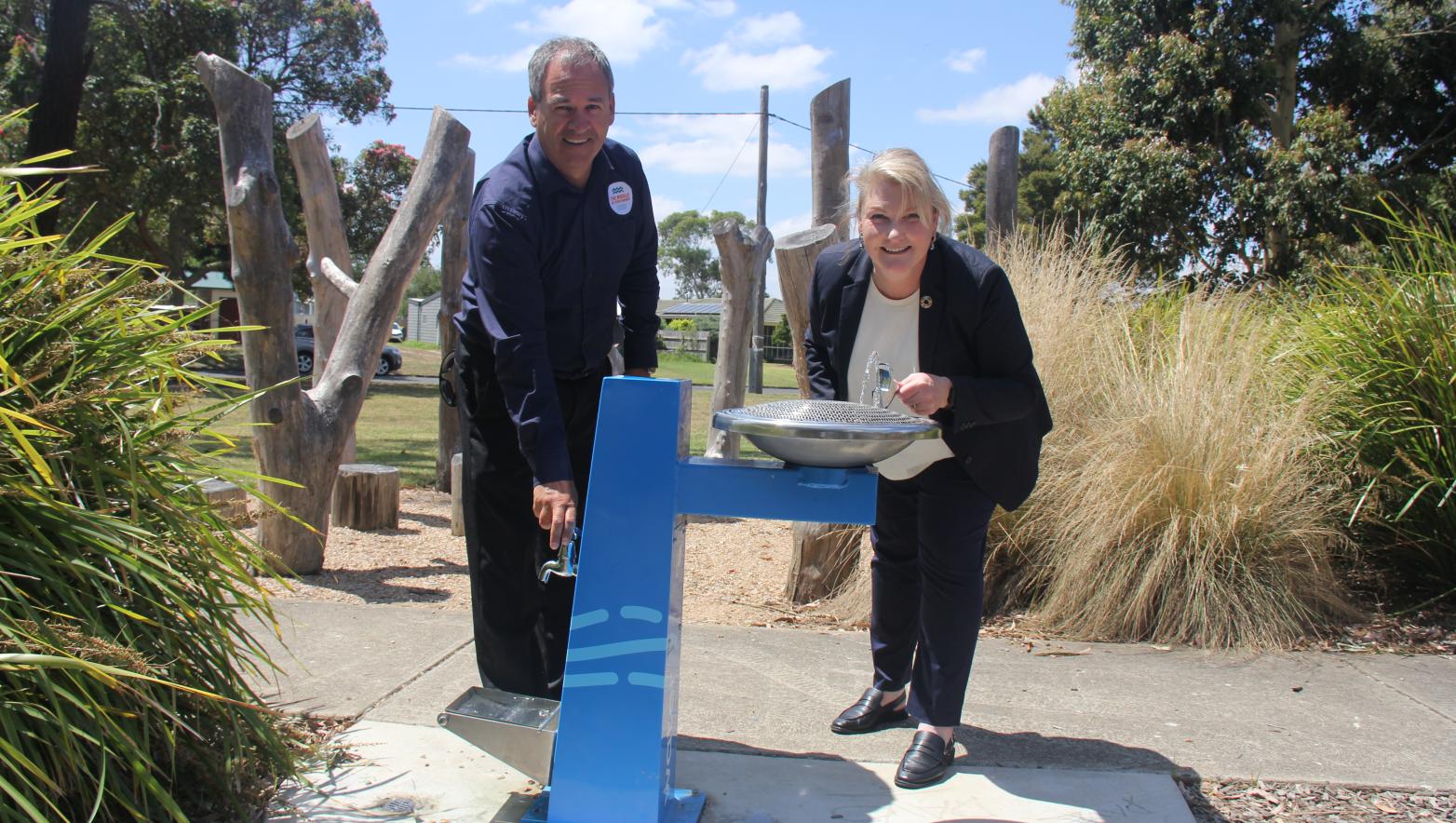 Man and woman stand at blue drinking fountain surrounded by shrubbery.