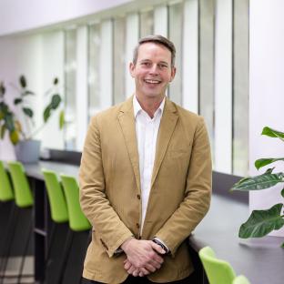 A tall man stands with hands folded smiling